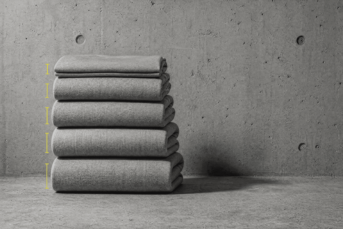 Five folded squares of grey sweatshirt cloth stacked in a column against concrete, each visibly thicker than the one below, with a measurement bracket beside each